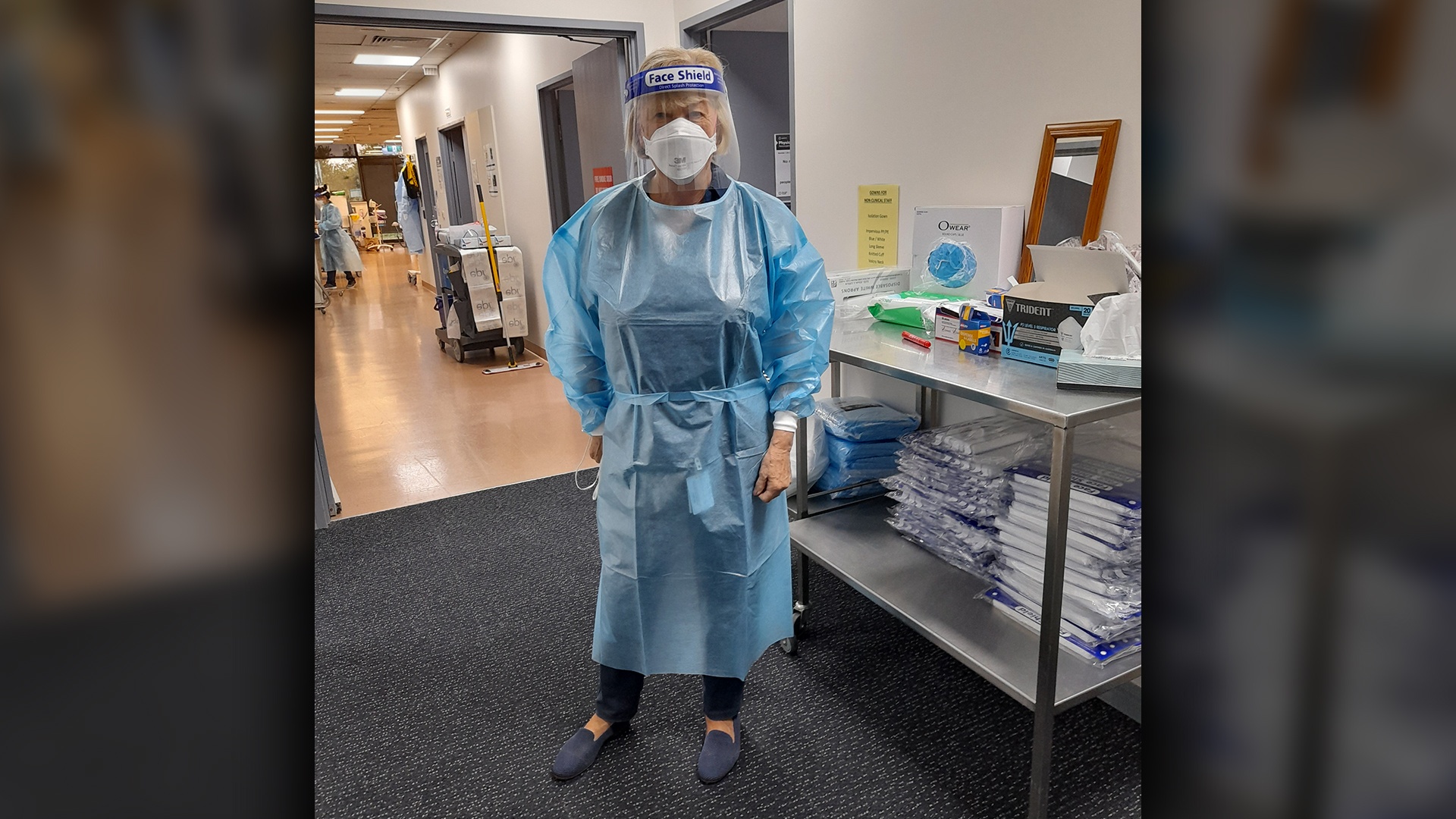 Volunteer Jenny Hill stands in a corridor, decked out head to toe in full PPE including a blue surgical gown, gloves, an N95 mask and a face shield.