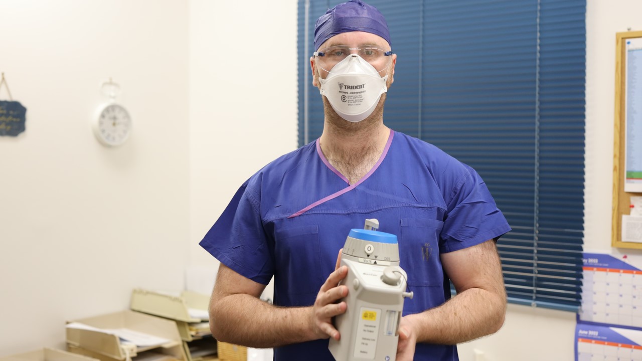 An anesthesiologist stands in a room holding a canister of the anesthetic agent desflurane. He wears blue scrubs, glasses and a white N95 mask.