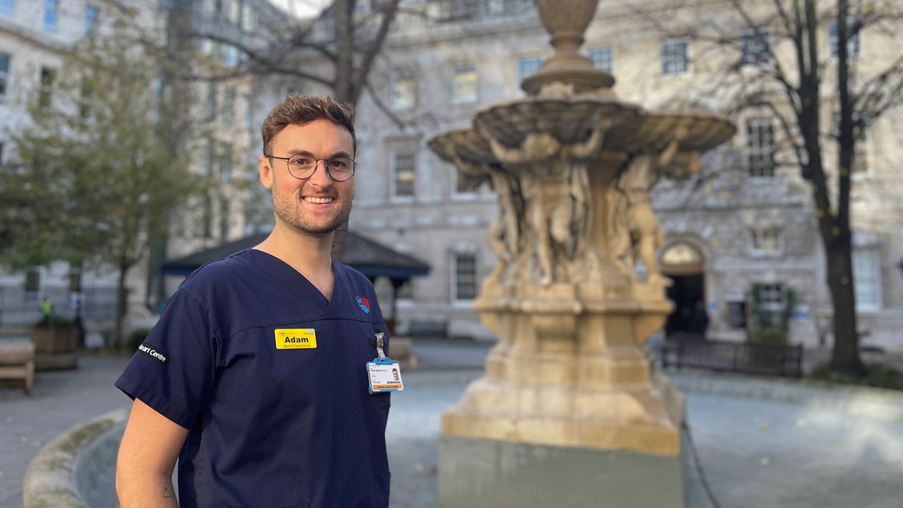Adam Damianopoulos stands in front of a fountain. He is wearing blue scrubs, a yellow name tag, glasses, and has short brown hair.