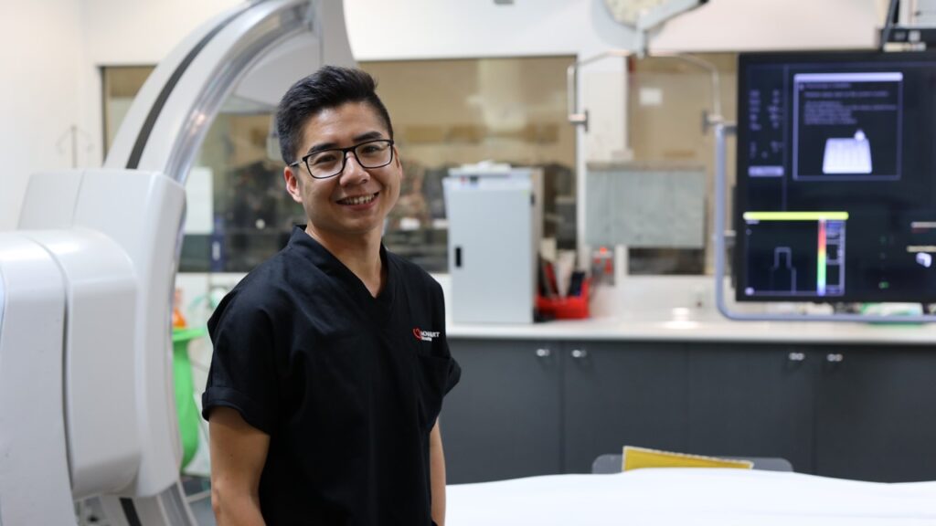 Trung Nguyen stands smiling inside a Cath Lab with machinery in the background. He wears dark navy scrubs, black square glasses and has black short hair.