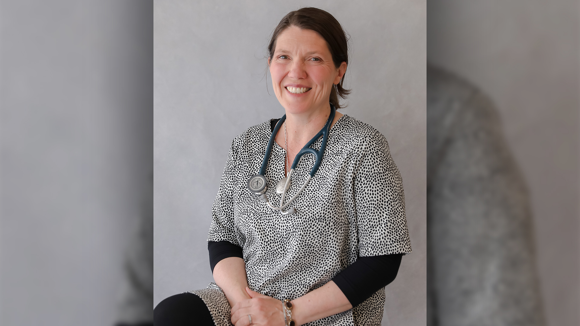 Director of Emergency Medicine Georgina Hayden sits with her hands clasped together on her lap and is smiling. She wears a black and white spotted shirt, a stethoscope and has her brown hair tied back in a low bun.
