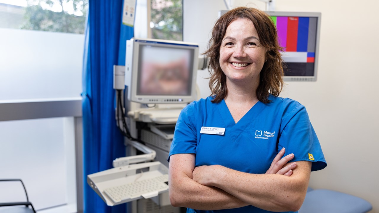 Speech Pathologist Claire Stanley stands in a clinical room with a bed and monitor in the background. She wears blue scrubs, has shoulder-length brown hair and is smiling with her arms crossed.