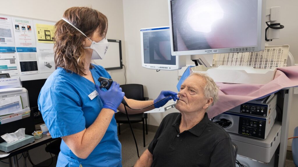 Speech Pathologist Claire Stanley performs a routine check of a patient's throat with an endoscope. The male patient is sitting wearing a black shirt, and Claire is wearing a blue shirt, gloves, a mask and is looking at him.