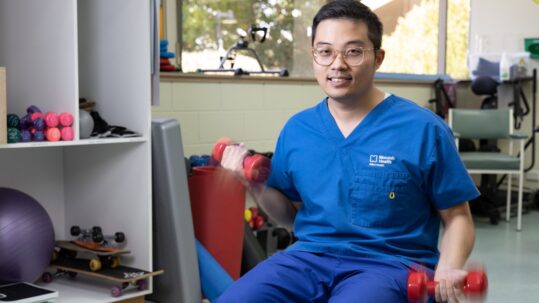Danny Nguyen sits in a room full of sporting equipment. He is lifting two red dumbbells with each of his hands and wears blue scrubs.