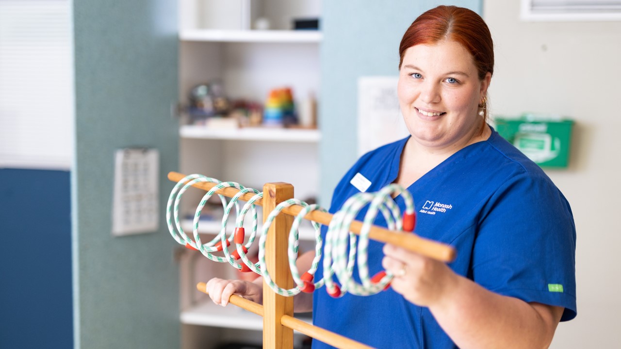 Eliza Beattie stands behind a wooden occupational therapy tool with rings on it, holding it with her hands and smiling. She wears blue scrubs and her auburn hair is tied back.