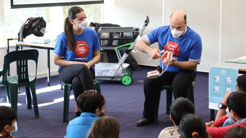 Two Dental Health Educators sit on chairs in a classroom full of children. One is using a giant model of teeth to show how you should be brushing.