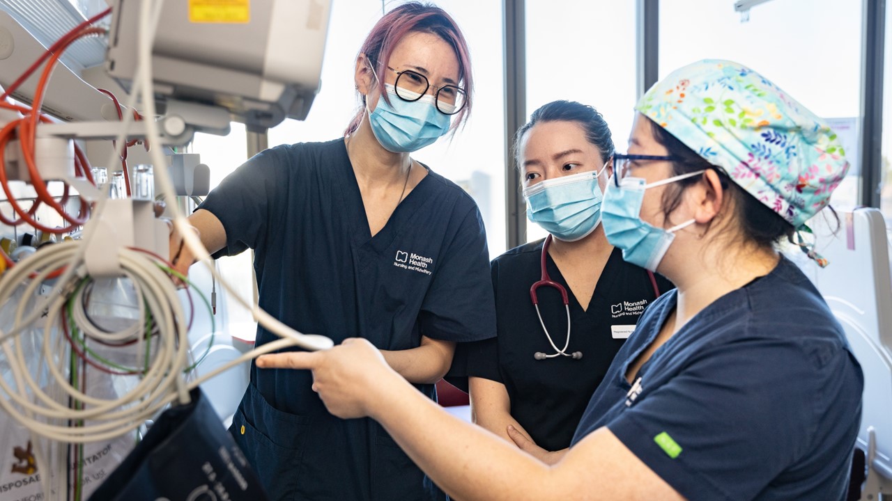 Registered Nurses Jo Tian, Tian Tan and Selby Li stand next to a monitor in discussion with each other. They all wear navy blue scrubs and masks.