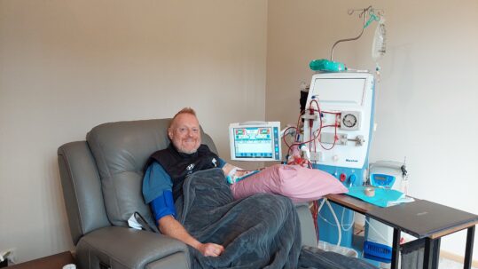 A man sitting on a chair with his left arm hooked up to tubes connected to a dialysis machine.