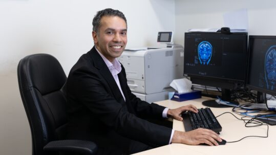 Dr Nevin John smiles at the camera, while facing two computer monitors showing a brain scan.