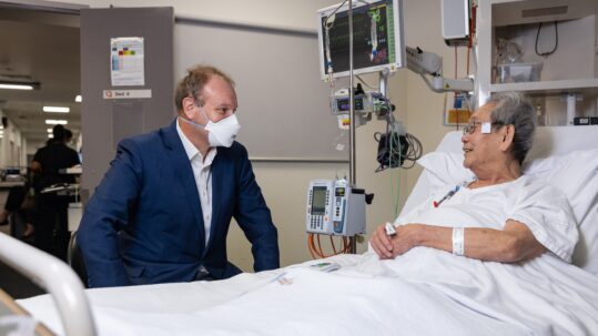 Professor Stephen Nicholls sits by a patient's bedside, interacting with him.