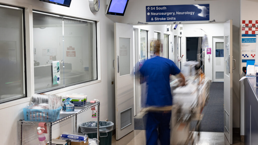 A blurry figure pushes a cart through a hallway with a sign above it saying "Neurosurgery, Neurosciences".