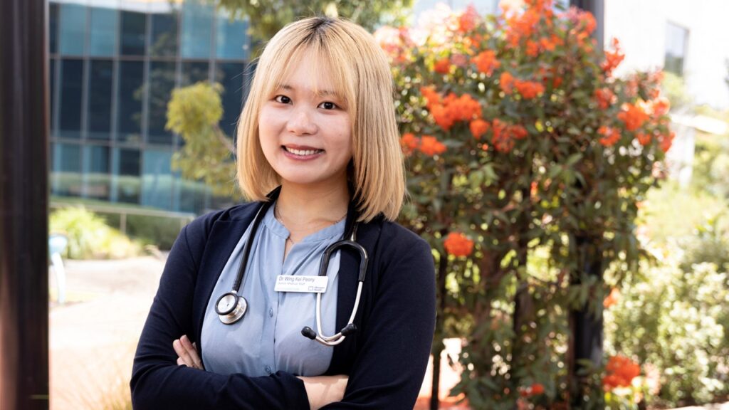 1920x1080px-WordPressFeaturedImage Peony Intern A young female doctor with a stethoscope draped around her neck stands in front of a flowering tree, smiling to camera