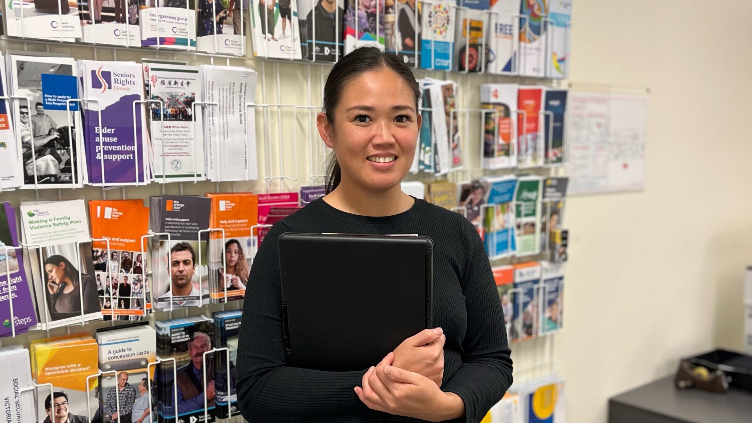 Graduate Social Worker Melissa Dang standing in front of a wall of information pamphlets holding a clipboard.