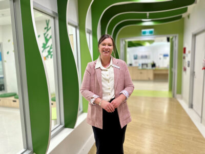 Tegan Dunmall, wearing a cream shirt and red and cream chequered jacket, smiles for a photo in a hospital walkway with bright green curved decor in the background.