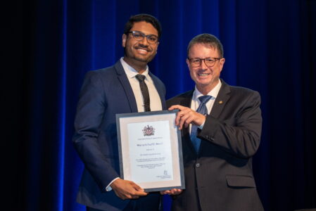 Two men in suits and ties smile for a photograph. Together the men are holding a framed award certificate.