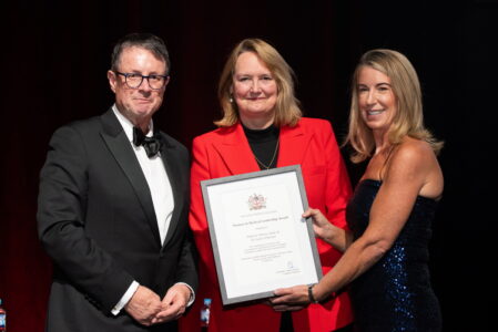 A woman in a bright red jacket smiles for a photograph alongside a man in a dinner jacket and a woman in a sequin dress holding a framed award certificate.