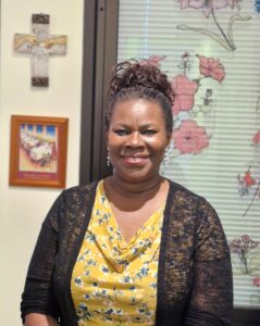 A black woman with a warm smile, on the wall behind her there are decorations, including flowers and a cross.