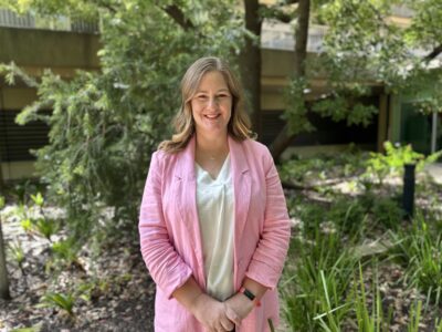 A smiling woman in a soft pink jacket and white t-shirt poses for a photo in a leafy garden.