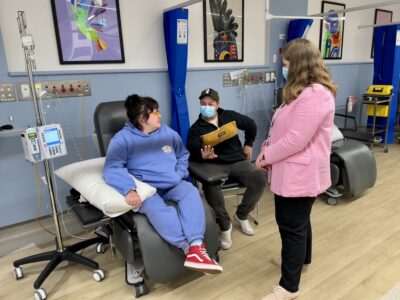 A woman receiving an infusion sits chatting with her partner and a nurse in a clinical setting.