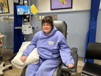 A woman in a light blue tracksuit sits in a recliner chair getting a transfusion.