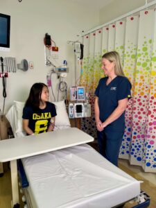 A nurse in navy blue scrubs stands at the bedside of an adolescent patient speaking with her.