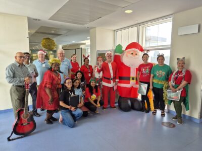 A man carrying a guitar and group of carolers wearing festive colours pose with an inflatable Santa for a photo.