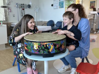 A young boy sitting on his mum's lap joyfully banging a large drum with a music therapist.
