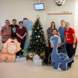 Staff wearing festive scrubs gathered around a Christmas tree surrounded by cardboard cutout dinosaurs.