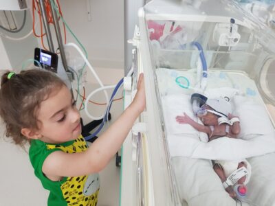 A 4-year-old girl peering through the side of a hospital bassinet at her premature baby sister inside.