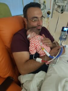 A dad sitting in a hospital chair cuddling his premature baby and holding the tubing connected to her.
