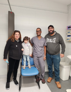 A doctor in medical scrubs in a consulting room smiles for a photo with a young girl and her parents.