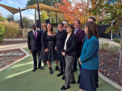 Around a dozen smiling people pose for a photo in an Autumnal Garden.