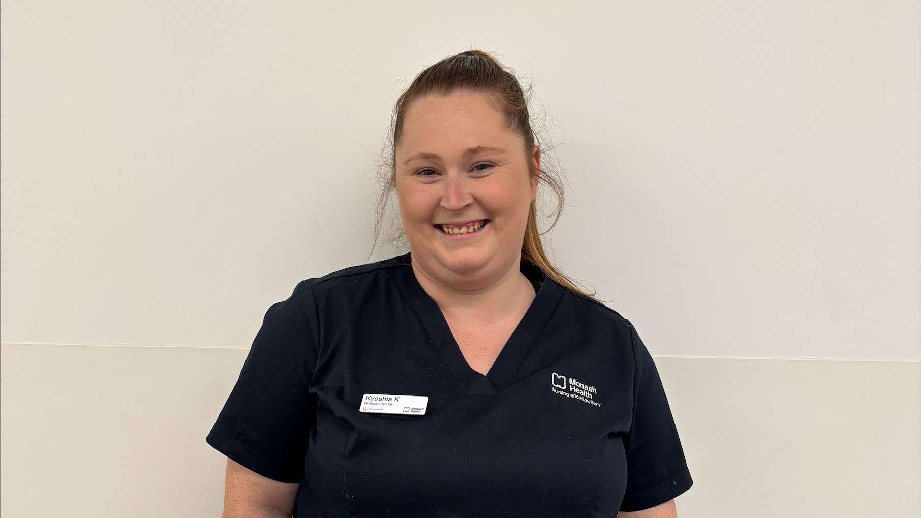A smiling woman with long brown hair in a ponytail. She is wearing navy blue medical scrubs.
