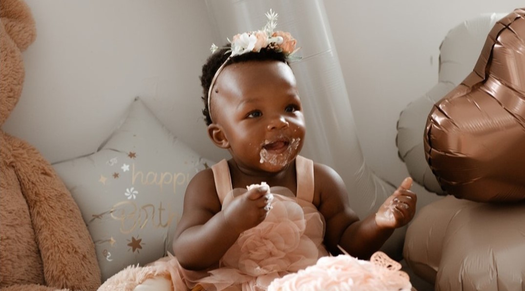 A smiling baby girl surrounded by balloons marking her first birthday. She has birthday cake around her mouth.