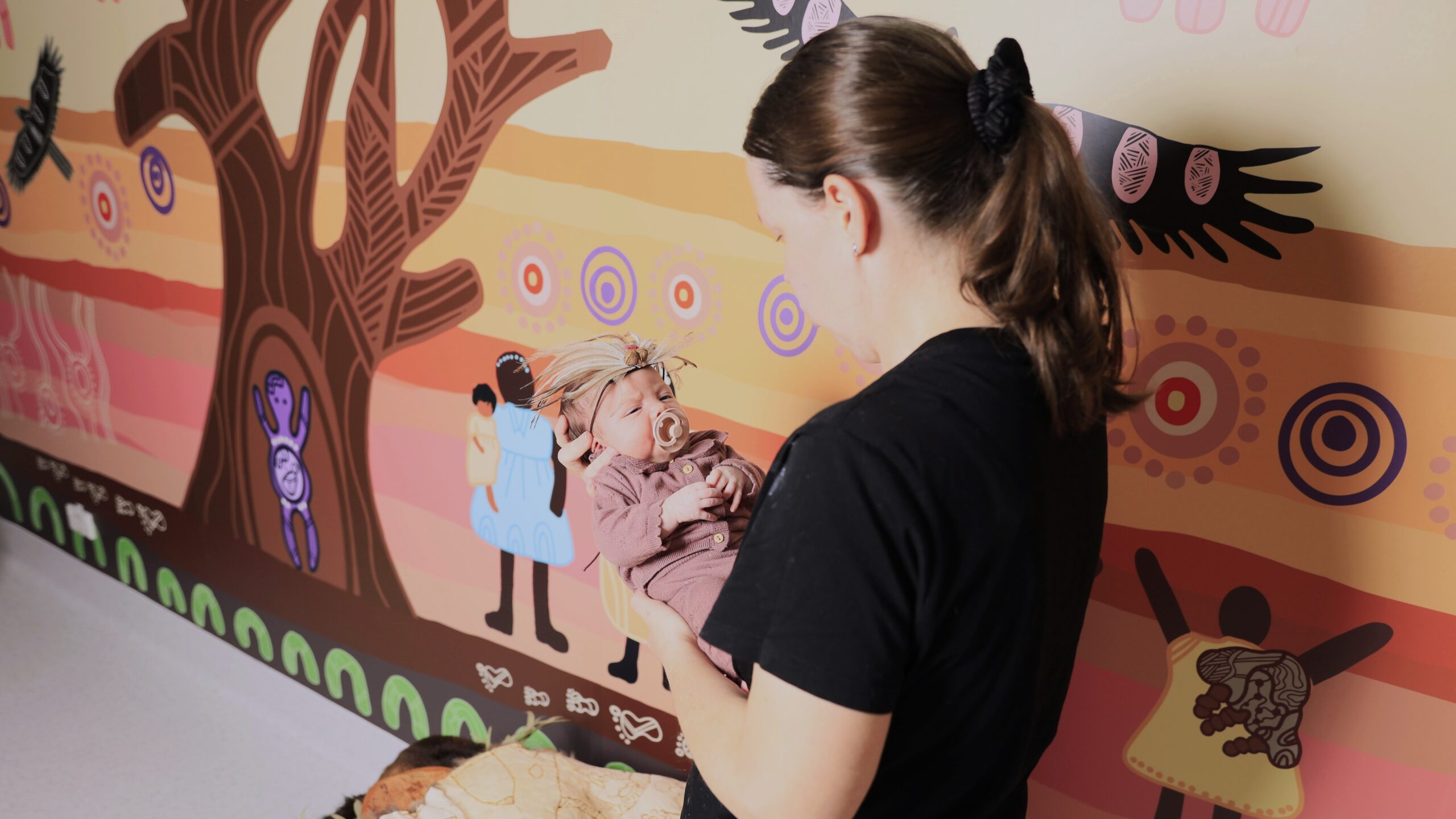 Photo of a baby being held by an Aboriginal Midwife Coordinator with the Bubup Dreaming artwork decal on a wall behind them.