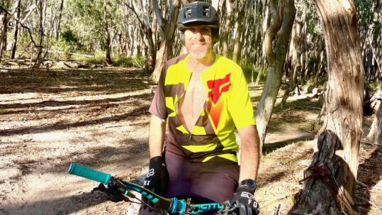 Alex sits on a mountain bike with his jersey unzipped to reveal a large scar on his chest.
