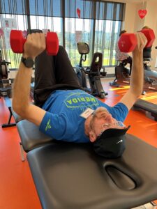 Alex lies on gym equipment raising two dumbells into the air.