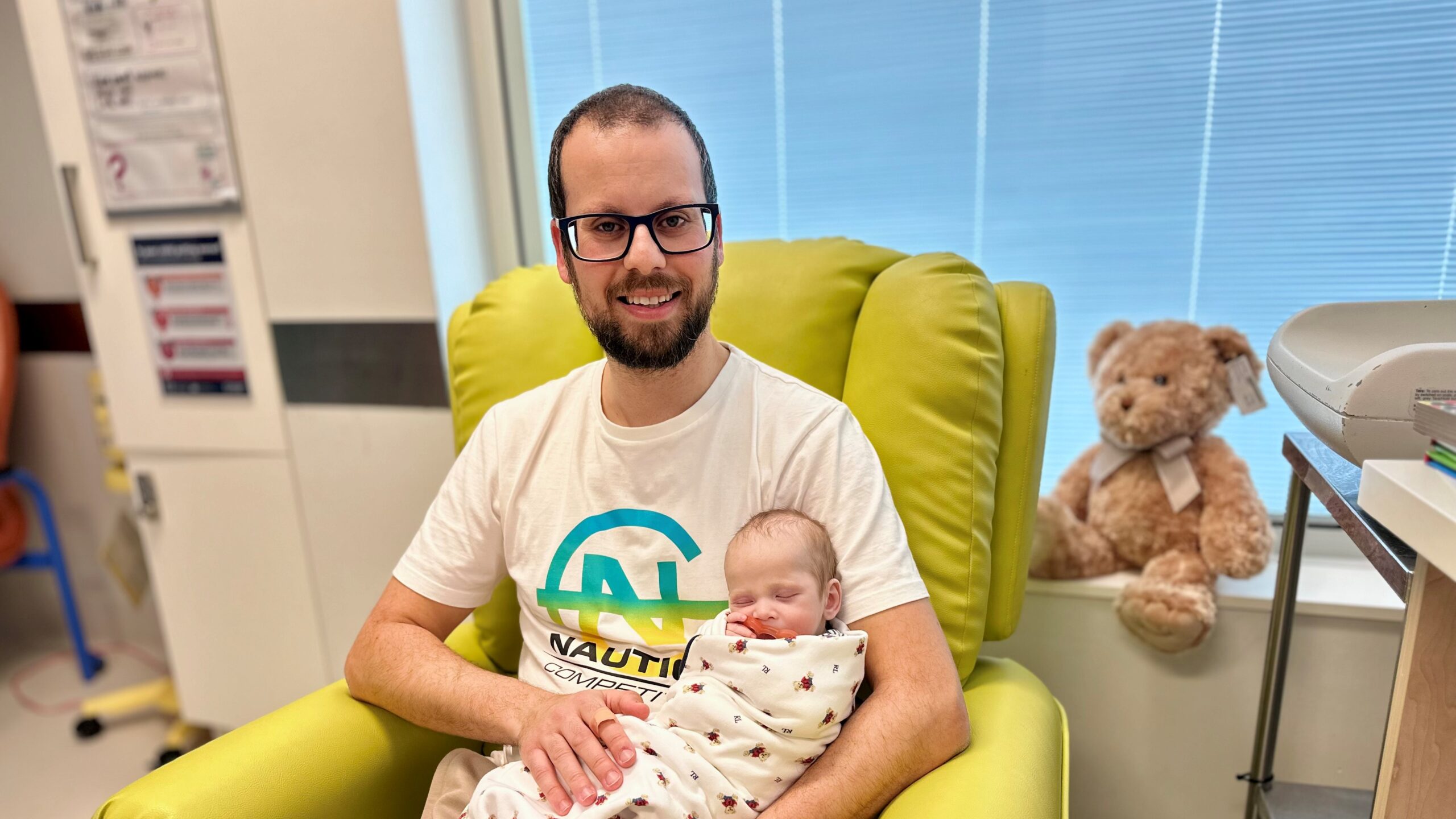 A dad sitting in a hospital armchair holding his baby son, who is wrapped in a blanket and sleeping soundly.