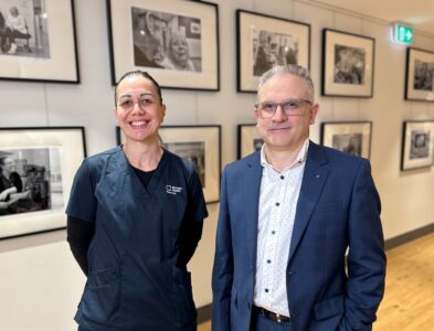 A smiling man and a woman pose for a photograph in front of a gallery of photos in a wide hospital corridor.