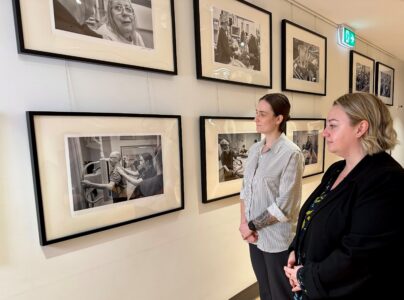 Two women looking at a collection of black and white framed photographs neatly arranged on a wall.