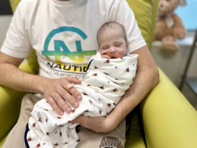 A swaddled baby with a dummy in his mouth sleeps soundly in his father's arm.