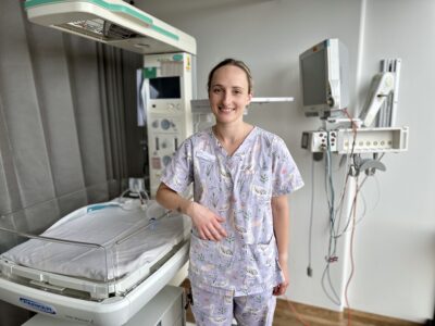 A smiling female midwife wearing medical scrubs stands by a bassinet in a clinical setting.