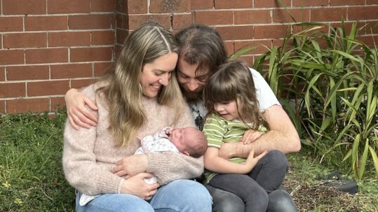 A woman cradling a baby in her arms. Her husband leans in close with his arm around her. Both look adoringly at the baby. A 4-year-old boy cuddles into them.