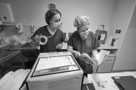 Two female transplant coordinators in medical scrubs, caps and masks checking paperwork as they seal a large, insulated box containing a donor kidney.