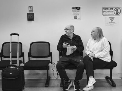 A man and a woman sit side-by-side in a waiting area. Beside them is a suitcase.