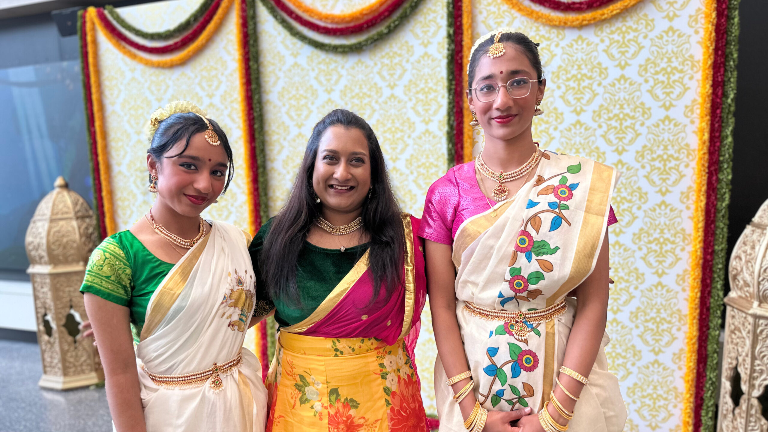 Three women wearing colourful sarees and gold jewelry standing in front of lanterns and floral wreaths at a Diwali celebration.