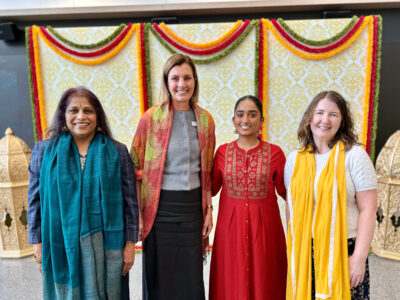 Four smiling women wearing colourful clothes, standing in front of gold lanterns and floral wreaths at a Diwali celebration.