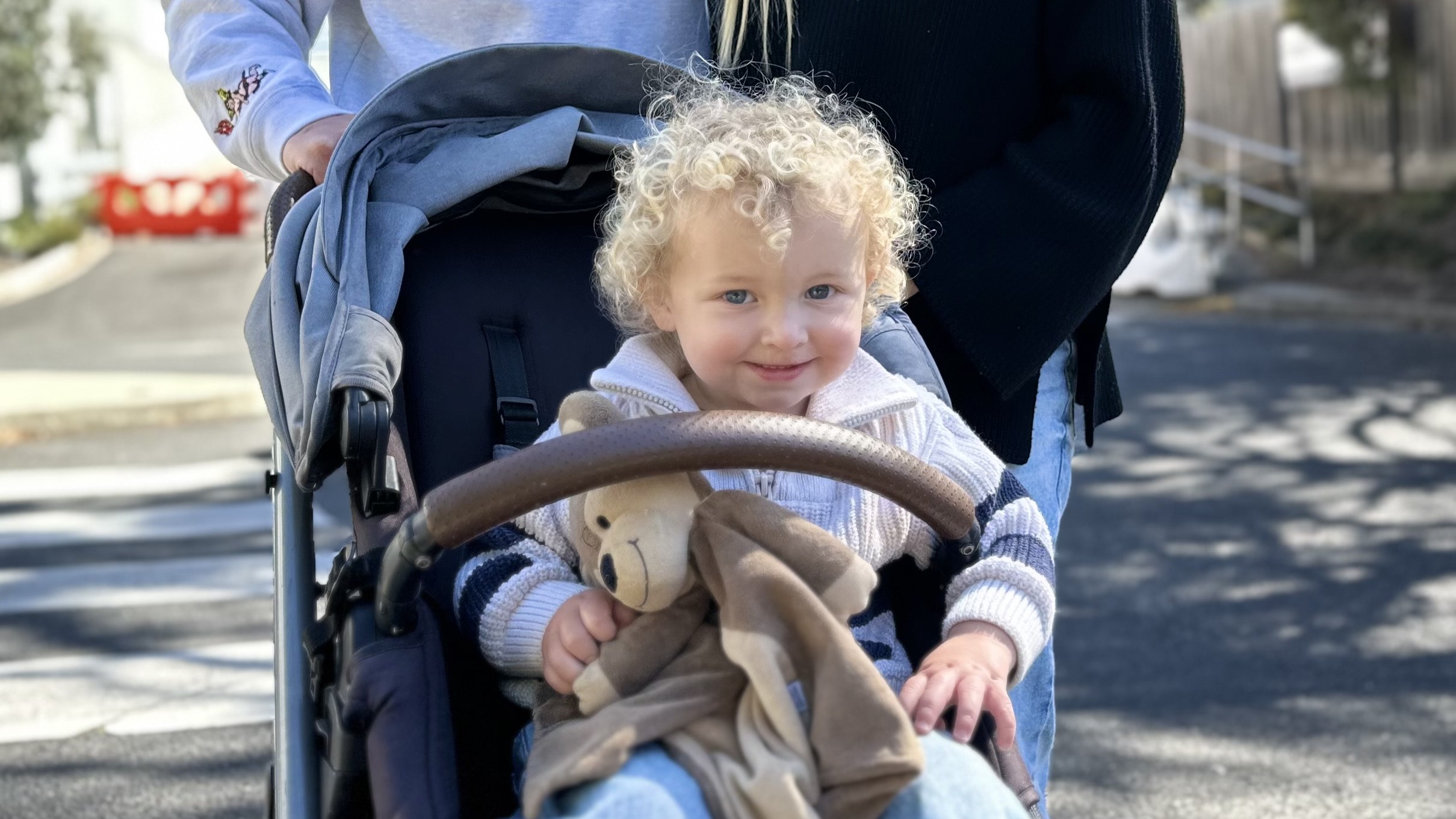 A smiling toddler with blond ringlets sitting in a pram holding a bear comforter.